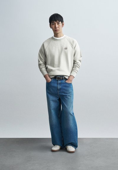 Ανοιχτό γκρι φούτερ με ανάγλυφη υφή, μπλε τζιν παντελόνι με φαρδυές παντελόνες και λευκά αθλητικά παπούτσια. Το φούτερ διαθέτει ένα μικρό λογότυπο στο στήθος.