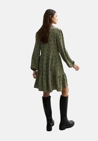 Robe verte à manches longues, à pois noirs, avec un ourlet en volants superposés, fabriquée en tissu texturé, portée avec des bottes noires jusqu'aux genoux.