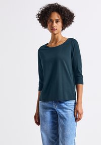 Mörk teal, långärmad topp med rund halsringning, tillverkad av mjukt material. I kombination med ljusblå, avslappnade jeans. Enkelt, oanimerat design.