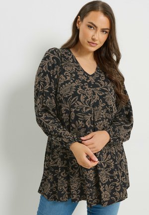 Kvinde iført en sort bluse med langærmede og beige blomsterprint, poserer med hænderne foldet og blå jeans mod en ensfarvet baggrund.