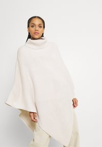 Poncho in morbido maglione beige con collo alto. Presenta una forma ampia e fluida e orli obliqui, offrendo una vestibilità rilassata.