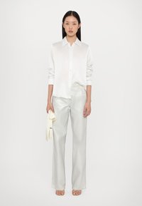 Chemise blanche en soie à boutons avec des manches longues, associée à un pantalon large en argent brillant. Le mannequin tient un accessoire de couleur claire.