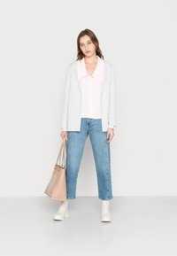 Blazer cinza claro sobre uma camisa de colarinho rosa, combinado com jeans azuis. O modelo carrega uma bolsa bege e usa botas brancas até o tornozelo.