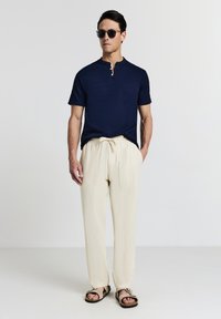 Maglietta henley a maniche corte blu navy con dettaglio a bottoni, abbinata a pantaloni beige con coulisse e sandali marroni, su uno sfondo neutro.