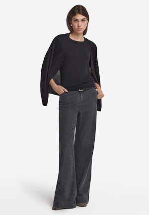Schwarzer Cardigan über einem schwarzen, figurbetonten Top, kombiniert mit dunkelgrauen, weit geschnittenen Jeans. Auffällige Details sind ein Gürtel und umgeschlagene Hosenabschlüsse.