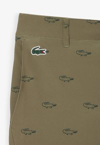 Khaki stof met groene krokodillogo's bedrukt en een groter groen en rood krokodillenembleem nabij de tailleband.