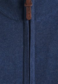 Námořnická modrá zapínací mikina se žebrovaným límcem a manžetami. Obsahuje hnědou koženou táhlo s embosovaným nápisem "JACKS". Měkká pletená struktura.