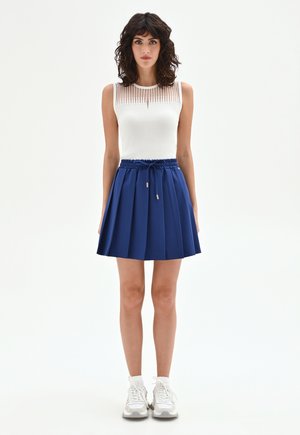 Kobieta o kręconych ciemnych włosach, ubrana w biały, bezrękawnik, niebieską plisowaną spódnicę ze ściągaczem, białe skarpetki i białe trampki, stojąca na białym tle.