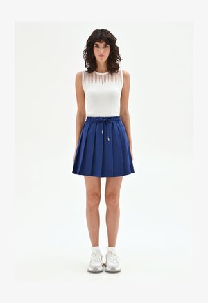 Femme aux cheveux bouclés et foncés portant un haut blanc sans manches, une jupe plissée bleue avec cordon de serrage, des chaussettes blanches et des baskets blanches, debout sur un fond blanc.