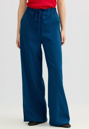 Mujer vistiendo pantalones azules de pierna ancha con cintura elástica y cordón, combinados con zapatos negros y top rojo, sobre un fondo liso.