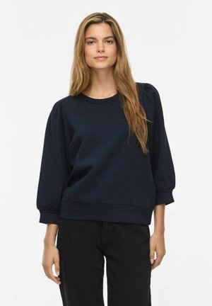 Jeune femme aux longs cheveux châtain clair portant un haut noir à manches longues à épaules bouffantes et un pantalon noir, se tenant devant un fond blanc uni.