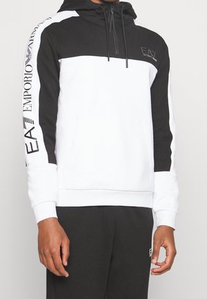 Mężczyzna noszący czarno-białą bluzę z kapturem EA7 Emporio Armani z napisem z logo na lewym rękawie i piersi.