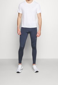 Homme portant une chemise blanche à manches courtes, des leggings de sport gris foncé et des chaussures de course blanches, se tenant devant un fond neutre.