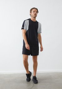 Schwarzes Sportshirt mit grauen Akzenten, kurze Ärmel, kombiniert mit schwarzen Shorts und schwarzen Sneakers, vor einem schlichten weißen Hintergrund.