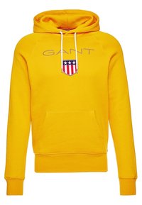 Sudadera amarilla de mezcla de algodón; cuenta con un bolsillo tipo canguro, cordones blancos y un logotipo en forma de escudo con detalles en rojo, blanco y azul.