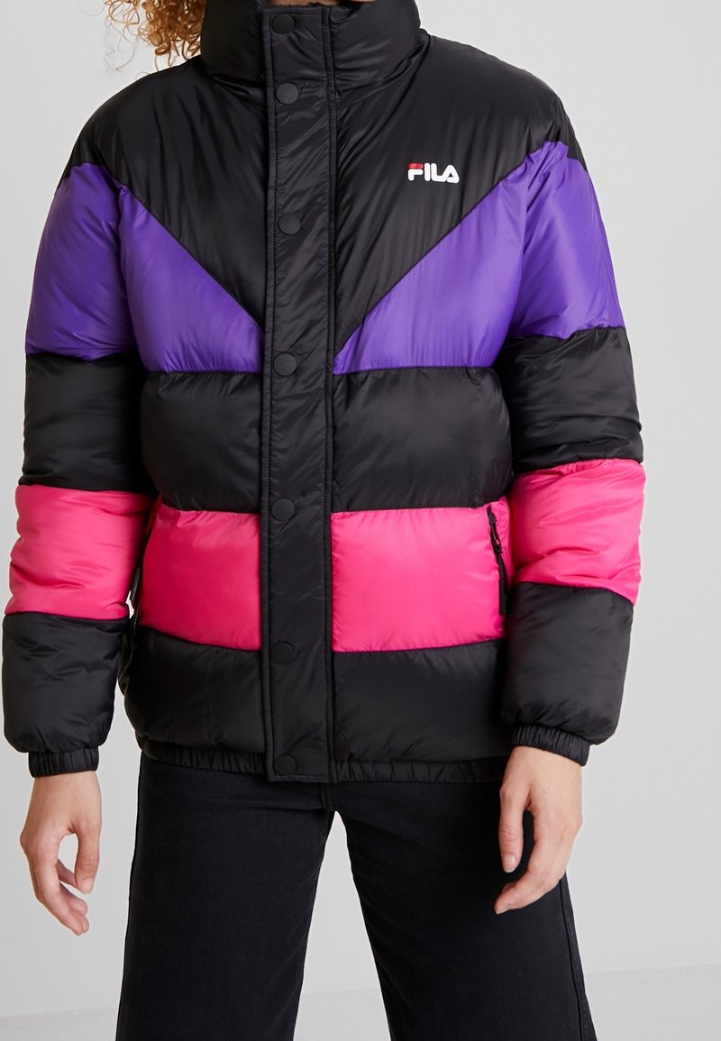 Personne portant une doudoune noire avec des blocs de couleur violet et rose, des boutons-pression à l’avant et le logo FILA sur la poitrine, associée à un pantalon noir.