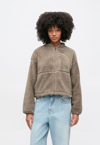 Žena s kučeravými vlasmi, oblečená v krátkej taupe fleecovej bunde a svetlomodrých džínsach s vysokým pásom, stojaca pred jednoduchým bielym pozadím.