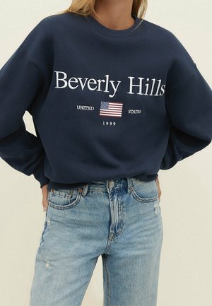 Vrouw in een marineblauwe sweatshirt met "Beverly Hills," een Amerikaanse vlag en de tekst "United States 1999," gecombineerd met lichtblauwe jeans.