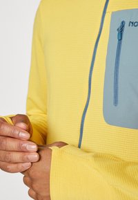 Yellow long-sleeve top with a textured finish, featuring a blue zippered chest pocket and contrasting trim around the cuffs.