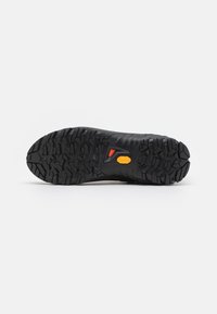 Svart tursko såle med dype, robuste spor mønstre, lett materiale og en lys gul Vibram-logo nær hælen.