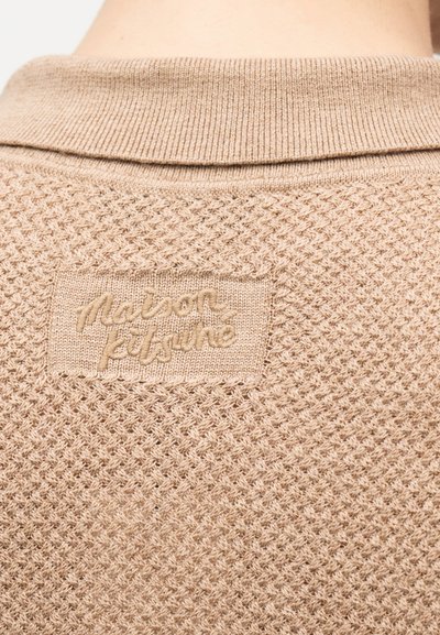 Κοντινό πλάνο μπεζ υφασμάτινου πουλόβερ με πλεκτή υφή, διπλωμένο γιακά και κεντημένο το «maison kitsuné» στην πλάτη.