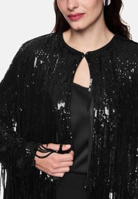 Black sequin cape with fringed design, featuring a round collar and keyhole closure. Smooth, shiny texture with intricate embellishments.