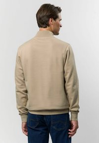 Béžová mikina s vysokým golierom, dlhými rukávmi a rebrovanými manžetami. Hladký povrch, voľný strih, kombinovaná s tmavomodrými džínsami.