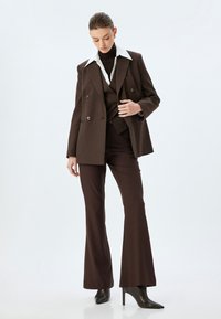 Dunkelbrauner maßgeschneiderter Anzug mit einem doppelreißigen Blazer, weißem Kragen und ausgestellten Hosen. Glattes Material und spitze High-Heel-Stiefel.