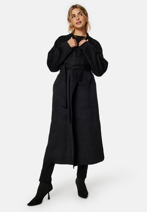 Black long coat made of soft fabric with a waist tie, featuring large pockets and wide sleeves. Worn over black pants and heeled shoes.