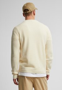 Suéter de canalé crema con mangas largas, presenta un escote redondo y un acabado texturizado, llevado encima de una camiseta blanca y combinado con pantalones claros.
