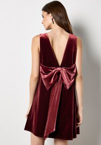 Bordowa sukienka z aksamitu z głębokim dekoltem na plecach, ozdobiona dużą kokardą w talii. Materiał ma miękką teksturę i połysk, a sylwetka jest rozkloszowana.