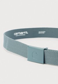 Ανοιχτό μπλε υφαντή υφασμάτινη ζώνη με λείο μεταλλικό αγκράφα, που φέρει ανάγλυφο καμπυλωτό έμβλημα, τοποθετημένη επίπεδη σε λευκή επιφάνεια.