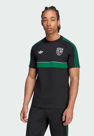 Młody mężczyzna z zaplecionymi włosami, ubrany w czarną sportową koszulkę Adidas z zielonymi paskami i logo RCL, stojący na jednolitym jasnym tle.