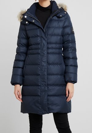 Manteau matelassé bleu marine avec fermeture éclair et boutons-pression, capuche bordée de fourrure, porté sur un col roulé noir et un pantalon, longueur mi-cuisse.