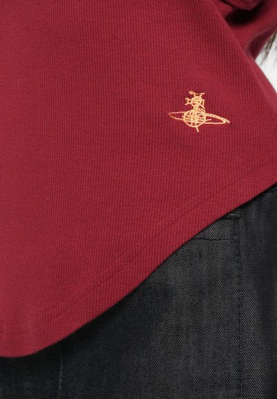 Burgundy színű pamut póló bordázott textúrával és ívelt szegéllyel, melyen arany hímzett logó található a bal oldalon, a szegély közelében.