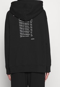 Svart hoodie med en stor huva, med vit text som listar datumen för modevisningar på baksidan. Mjuk tyg, avslappnad passform och ribbad nederkant.