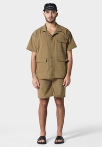 Chemise à manches courtes tan avec deux poches, assortie à un short. Casquette noire avec texte blanc et sandales à glisser noires. Texture de tissu lisse.
