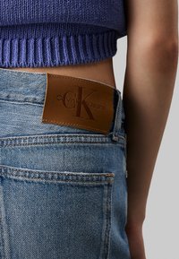 Vaqueros de mezclilla azul con parche de cuero marrón de Calvin Klein Jeans. Las características incluyen costuras visibles y un diseño clásico de 5 bolsillos.