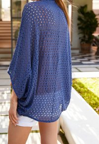 Blaues offenes Strickjacke mit lockeren Ärmeln, ausgestattet mit einem strukturierten Design und einer entspannten Passform, präsentiert vor einer hellen Outdoor-Kulisse.