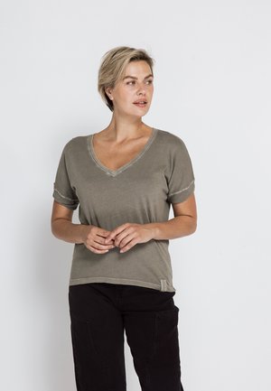 Frau mit kurzen blonden Haaren, die ein graues T-Shirt mit V-Ausschnitt und kurzen Ärmeln sowie schwarze Hose trägt, steht und schaut nach rechts.