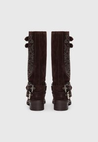 Stivali alti fino al ginocchio in suede marrone con motivi decorativi, fibbie in metallo argentato alla caviglia e fiocchi decorativi in cima.