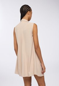 Abito beige senza maniche con colletto. Tessuto morbido e leggero, vestibilità ampia, presenta una piega sul retro per un ulteriore movimento e comfort.