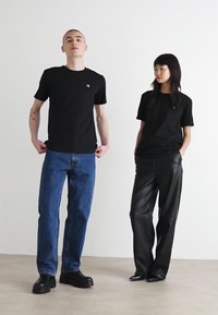 Dos individuos llevan camisetas negras. Uno tiene jeans de mezclilla azules; el otro tiene pantalones de cuero negros. Ambos llevan calzado oscuro.