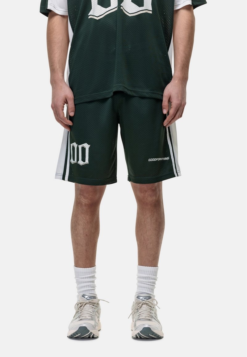 Zielone siatkowe szorty sportowe z białymi akcentami, wyraźnym nadrukiem numeru i logo. Dobrane do szarych sneakersów i prążkowanych skarpet.