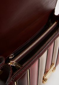 Interior de un bolso de cuero con un forro burdeos; cuenta con un compartimento con cremallera con herrajes dorados y patrones de rayas alternos en beige y rosa.