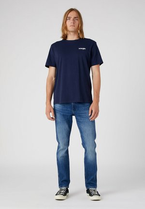 Jong persoon met schouderlang haar, een marineblauw Wrangler T-shirt, blauwe spijkerbroek en zwart-witte sneakers, staand tegen een effen achtergrond.