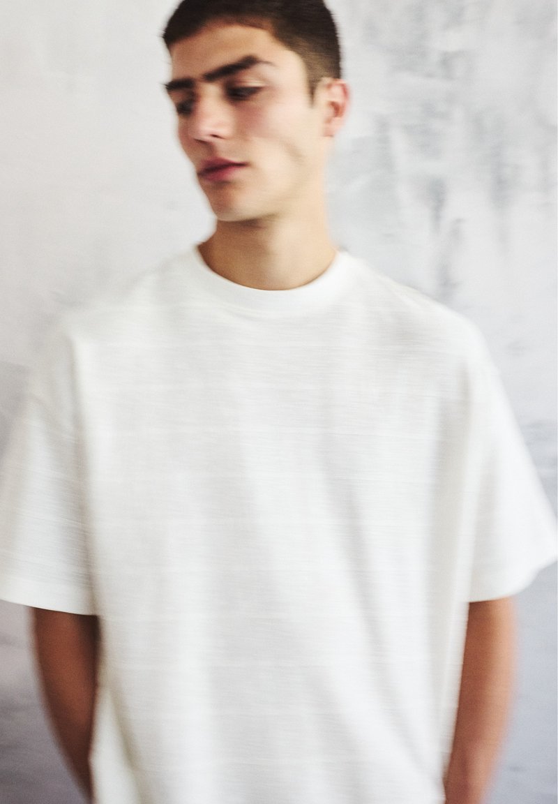 Young man in white t-shirt standing against a light gray textured wall, looking slightly downward to his left.