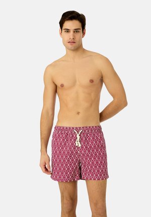 Homme torse nu aux cheveux foncés portant un short de bain rouge à motifs de losanges blancs et un cordon blanc, debout devant un fond uni.