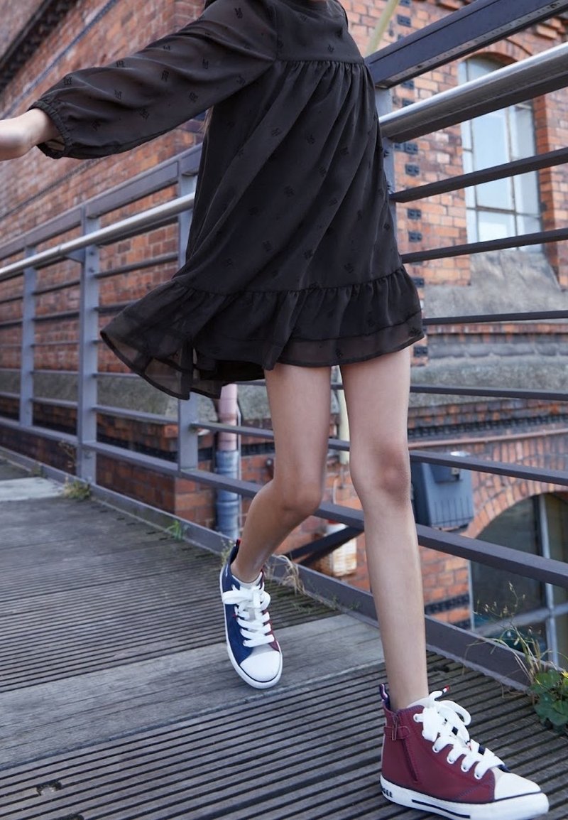 Vysoké tenisky v námořnické modré a burgundské plátno s bílou gumovou podrážkou, doplněné bílými tkaničkami a akcentovými detaily. Proužkované černé šaty dodávají pohyb.