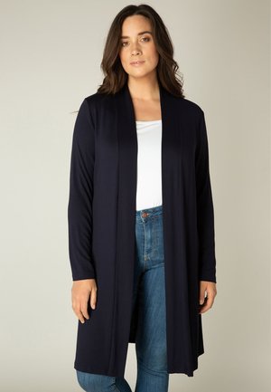 Vrouw draagt een lange marineblauwe open voorkant cardigan over een wit topje en blauwe spijkerbroek, staand tegen een effen neutrale achtergrond.